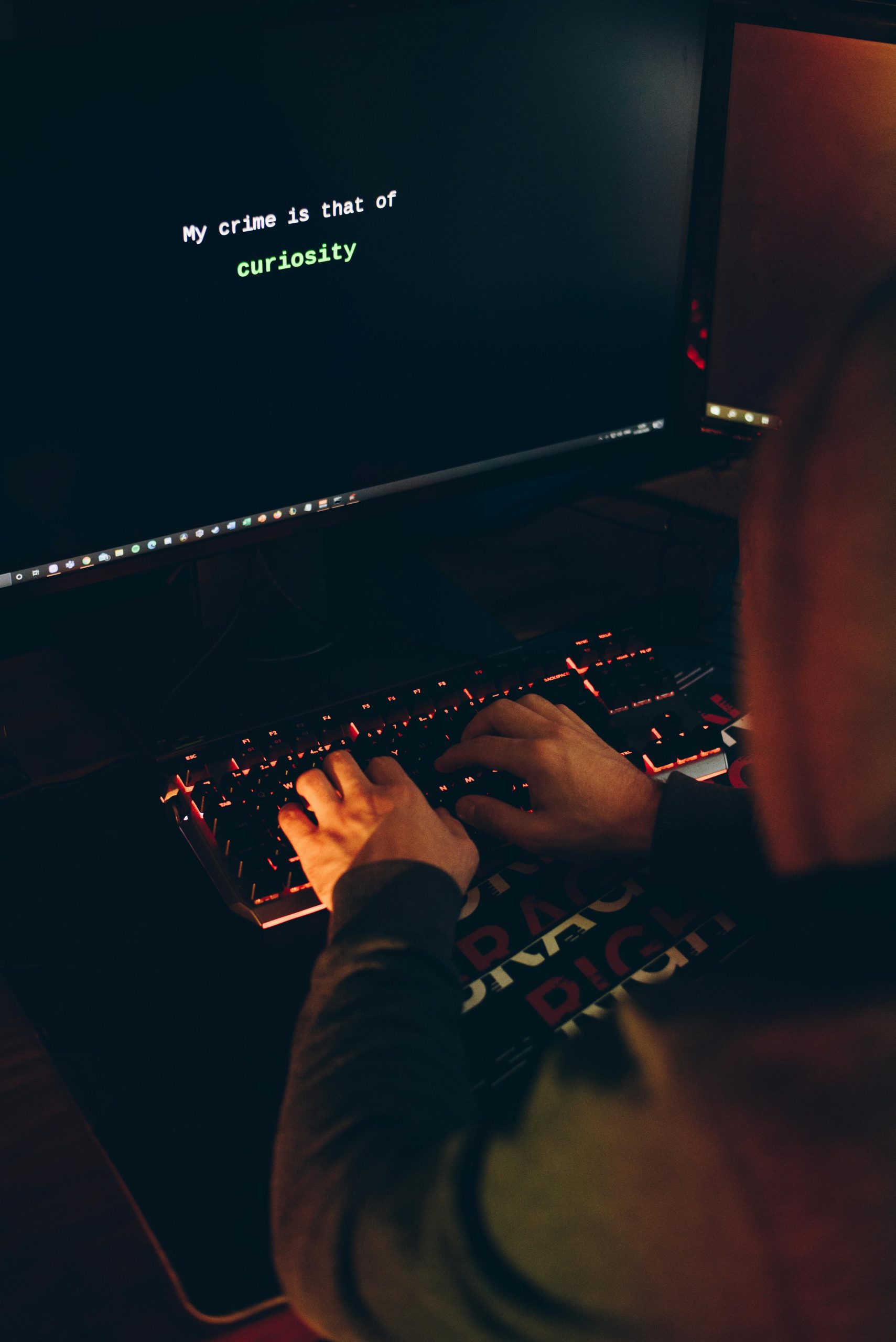 A person in a hoodie typing on a keyboard with a computer screen displaying a curiosity message.