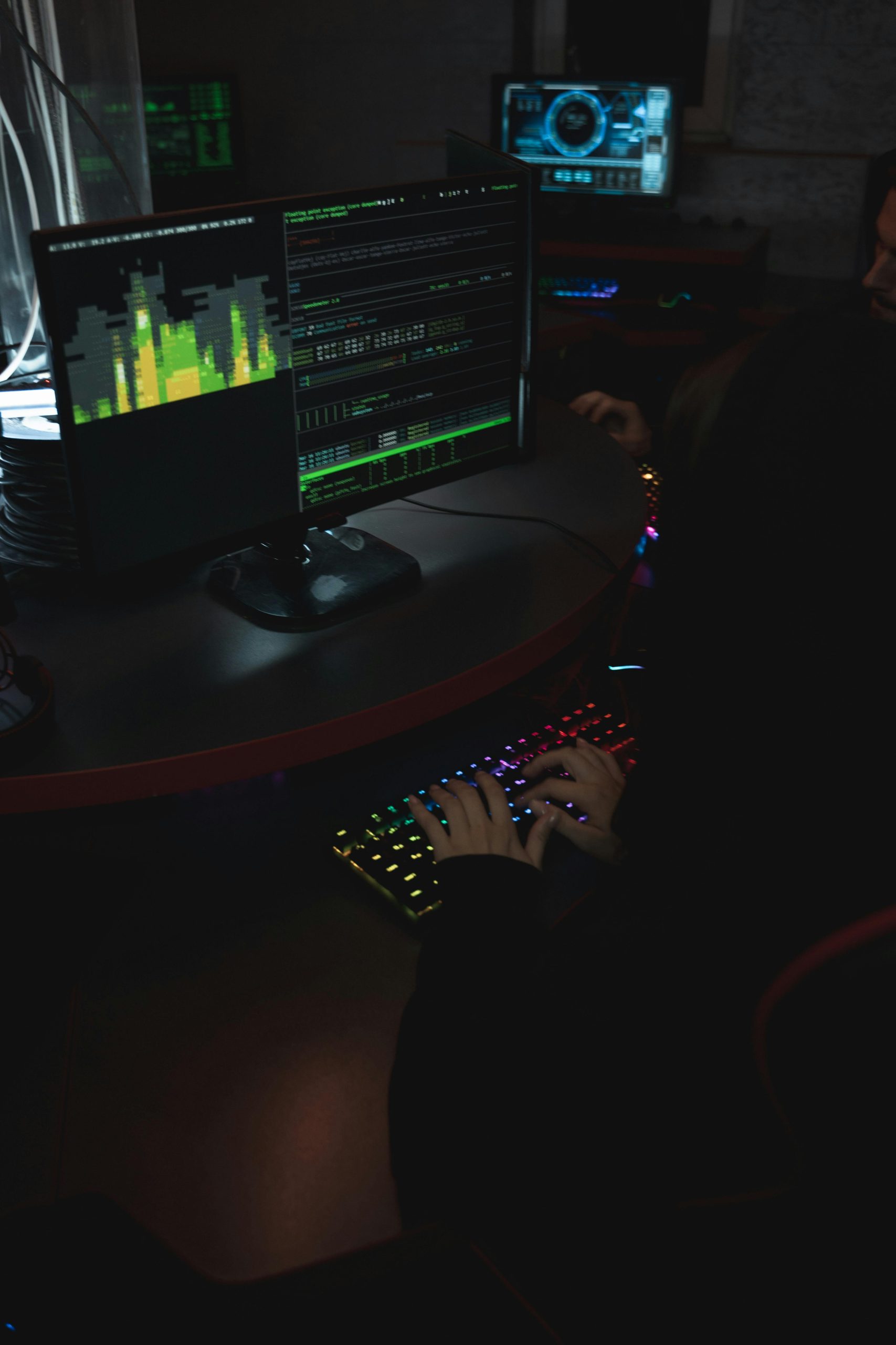 Person typing on RGB keyboard in a dark room with coding screens.