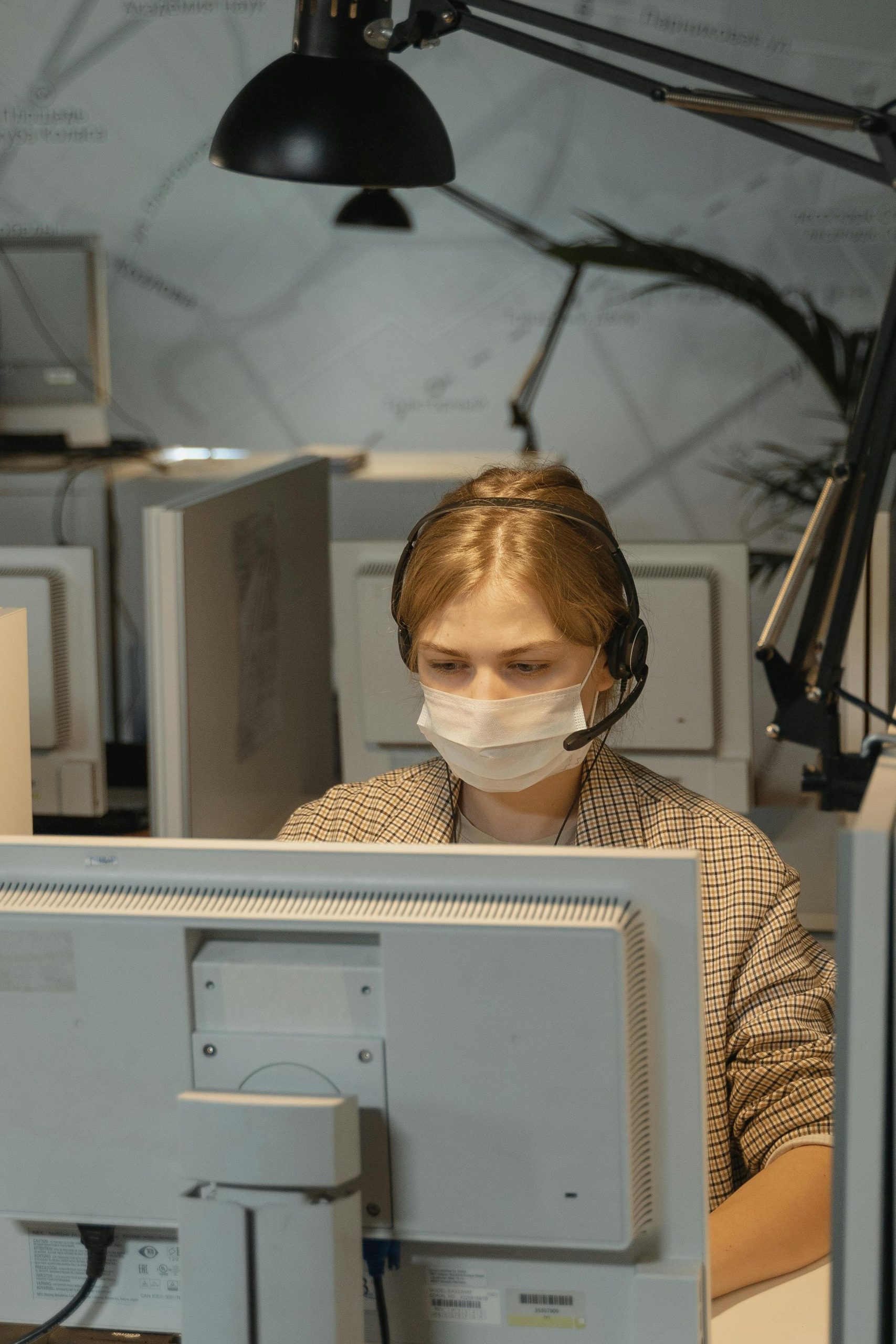 Call center employee wearing mask and headset, focused on computer work.